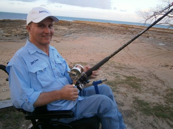 Hands-On adaptive fishing rod holder for under wheelchair seat - man in wheelchair on beach hand cranked